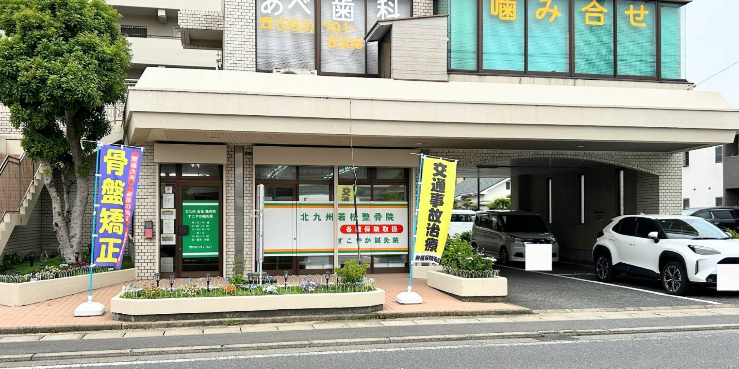 北九州市若松区の「すこやか鍼灸整骨院」の外観。入口には「骨盤矯正」「交通事故治療」ののぼり旗が掲げられており、窓には各種保険取扱の案内が表示されている。
